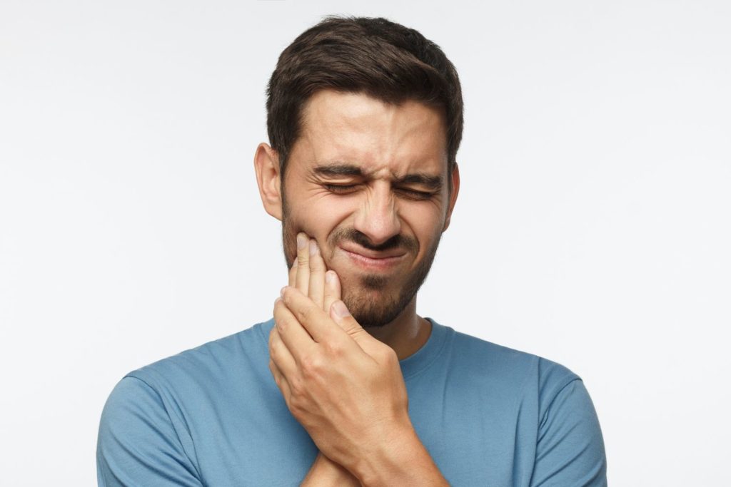 young guy isolated on gray background, his face distorted and hands on his jaw in pain TMJ restorative dentistry dentist in Monroe North Carolina